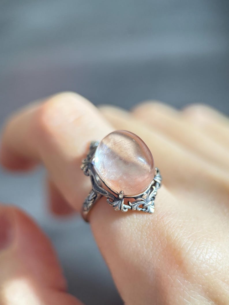 Op de afbeelding: Een zilveren ring met een ovale, doorschijnende, perzik-kleurige edelsteen. De ring heeft een sierlijk filigraan ontwerp met blad- en kruldetails rond de steen. De ring wordt aan een vinger gedragen.