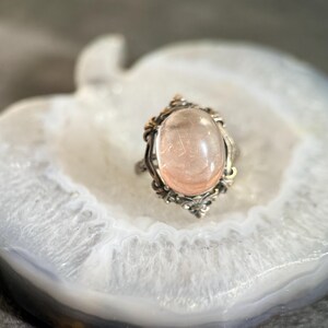 Op de afbeelding: Een ring in vintage-stijl met een ovale, lichtroze edelsteen gezet in een zilverkleurig metalen frame. Het frame heeft sierlijke details met gouden accenten, wat een antieke uitstraling cre&euml;ert. De ring ligt op een wit-grijze ondergrond.