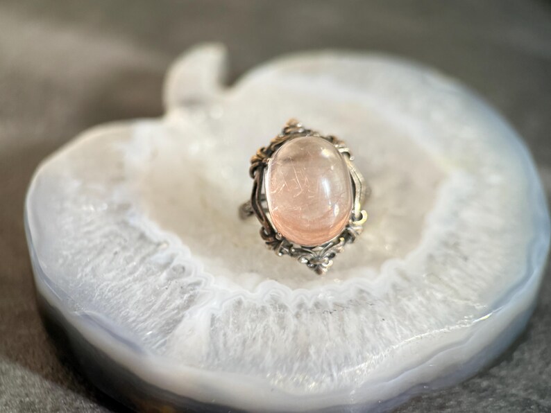 Op de afbeelding: Een ring in vintage-stijl met een ovale, lichtroze edelsteen gezet in een zilverkleurig metalen frame. Het frame heeft sierlijke details met gouden accenten, wat een antieke uitstraling cre&euml;ert. De ring ligt op een wit-grijze ondergrond.