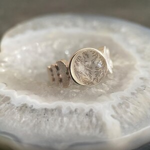 May include: A silver ring with a clear round stone featuring a delicate fern pattern inside. The ring is resting on a white agate surface.