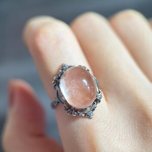 Op de afbeelding: Een zilveren ring met een ovale, perzikkleurige edelsteen. De ring heeft een sierlijk filigraanontwerp rond de steen, met gedetailleerde krullen. De edelsteen is glad en doorschijnend, gezet in een vintage-stijl setting.