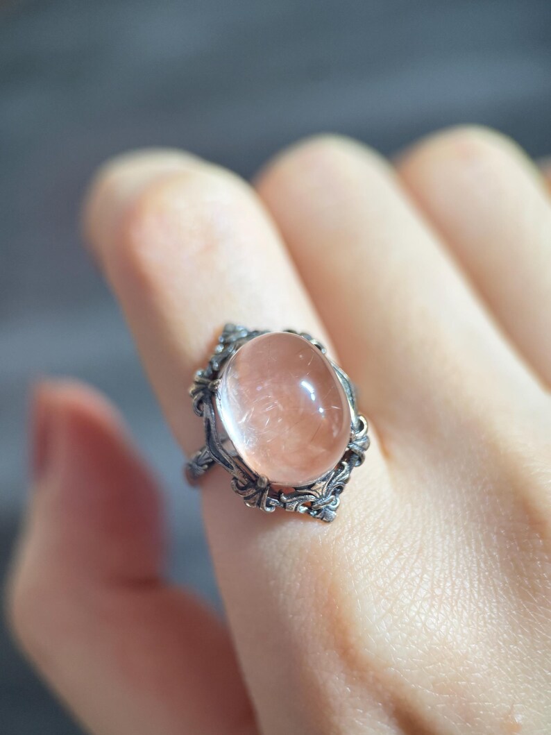 Op de afbeelding: Een zilveren ring met een ovale, perzikkleurige edelsteen. De ring heeft een sierlijk filigraanontwerp rond de steen, met gedetailleerde krullen. De edelsteen is glad en doorschijnend, gezet in een vintage-stijl setting.