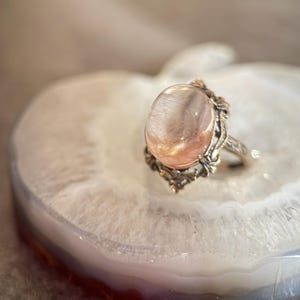 Op de afbeelding: Een vintage ring met een grote, ovale, doorschijnende roze edelsteen. De ring heeft een sierlijke, gedetailleerde zilverkleurige zetting met decoratieve versieringen. De ring wordt gepresenteerd op een witte en grijze agaat schijf.