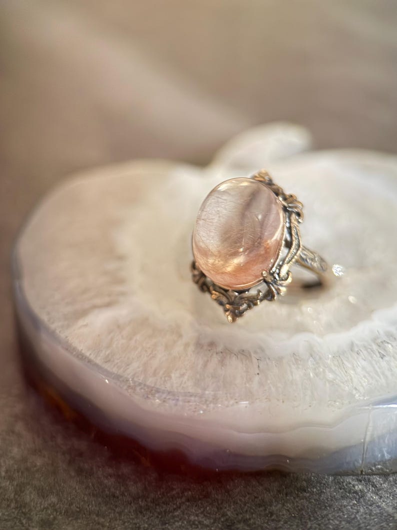 Op de afbeelding: Een vintage ring met een grote, ovale, doorschijnende roze edelsteen. De ring heeft een sierlijke, gedetailleerde zilverkleurige zetting met decoratieve versieringen. De ring wordt gepresenteerd op een witte en grijze agaat schijf.