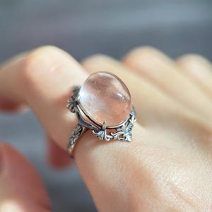 Op de afbeelding: Een zilveren ring met een grote, ovale, doorschijnende roze edelsteen. De ring heeft een sierlijk, bloemachtig ontwerp rond de edelsteen en op de band. De ring wordt aan een vinger getoond.
