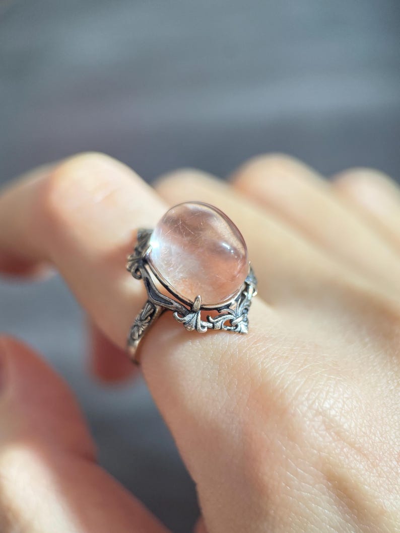 Op de afbeelding: Een zilveren ring met een grote, ovale, doorschijnende roze edelsteen. De ring heeft een sierlijk, bloemachtig ontwerp rond de edelsteen en op de band. De ring wordt aan een vinger getoond.