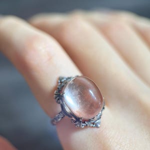 Op de afbeelding: Een zilverkleurige ring met een ovale, perzik-kleurige edelsteen. De ring heeft een sierlijk filigraanontwerp rond de steen en op de band. De edelsteen heeft interne insluitsels en een gepolijst oppervlak.