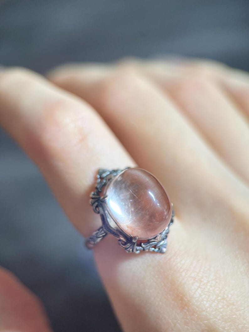 Op de afbeelding: Een zilverkleurige ring met een ovale, perzik-kleurige edelsteen. De ring heeft een sierlijk filigraanontwerp rond de steen en op de band. De edelsteen heeft interne insluitsels en een gepolijst oppervlak.