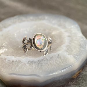 Puede incluir: Un anillo de plata con un delicado diseño floral que presenta una piedra redonda de nácar de abulón iridiscente.