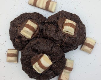 May include: Four chocolate chip cookies with white chocolate bars on top. The cookies are on a white surface.