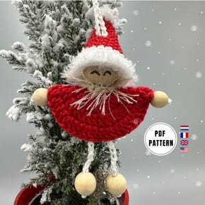 Op de afbeelding: Gehaakte kerstversiering van een kerstman met een rode muts, witte baard en houten kralenaccenten. De versiering hangt aan een kleine besneeuwde kerstboom. De tekst "PDF PATTERN" is zichtbaar op de afbeelding.