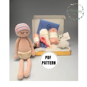 Op de afbeelding: Een haak-amigurumi poppenset met roze en wit garen, een blauw-wit gestreepte muts en een PDF-patroon. De set bevat ook een bedankkaart.