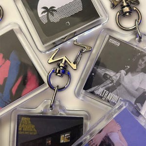 May include: A selection of clear acrylic keyrings, each containing album art inserts. The keyrings have silver-coloured metal fixings, including key rings and star-shaped connectors. The album art displays a range of colours and designs, including monochrome images and text.
