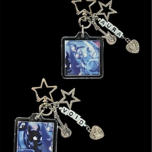 Puede incluir: Dos llaveros con una imagen cuadrada de un personaje de dibujos animados azul. Uno tiene la palabra "AURA" en cuentas blancas, el otro "VOID". Cada llavero tiene un cierre en forma de estrella, un dije de guitarra y un dije de corazón.