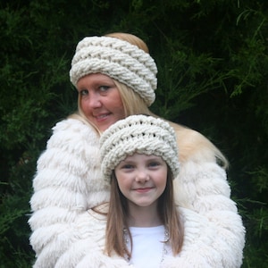 Peut inclure: Deux personnes portant des bandeaux crochetés crème assortis et des vestes en fausse fourrure crème. Les bandeaux ont un motif épais et texturé. L'arrière-plan est un buisson vert.