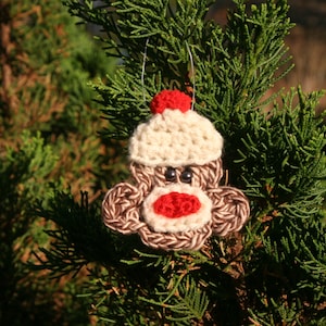 May include: A crocheted monkey ornament with a red hat, brown fur, and black eyes. The ornament is hanging on a green pine tree branch.