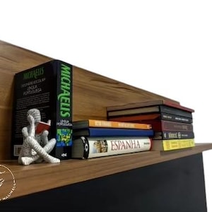 K&ouml;nnte beinhalten: Ein Holzregal mit einer Vielzahl von B&uuml;chern, die darauf gestapelt sind. Die B&uuml;cher sind in verschiedenen Farben und Gr&ouml;&szlig;en, und einige haben sichtbare Titel. Eine kleine silberne Figur eines lesenden Menschen steht auf dem Regal.