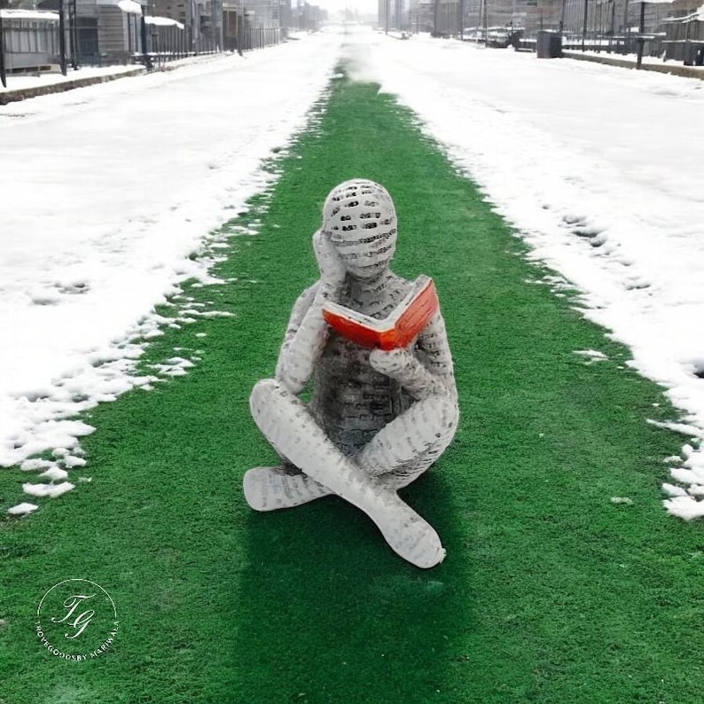 K&ouml;nnte beinhalten: Eine wei&szlig;e, papierartige Skulptur einer Person, die auf einem gr&uuml;nen k&uuml;nstlichen Rasenweg sitzt und ein rotes Buch liest. Der Weg ist von Schnee umgeben.