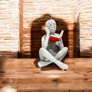K&ouml;nnte beinhalten: Eine wei&szlig;e, abstrakte Skulptur einer Person, die mit einem roten Buch in den H&auml;nden sitzt. Die Skulptur steht auf einer Holzoberfl&auml;che vor einer Backsteinwand.