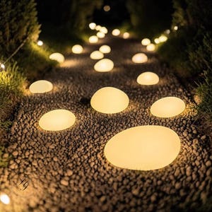 May include: A gravel pathway illuminated by glowing, oval-shaped lights, resembling stones. The lights are spaced along the path, creating a warm ambiance. Green foliage lines the sides, with additional small lights in the background, creating a garden path.