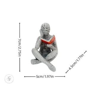 K&ouml;nnte beinhalten: Eine wei&szlig;e, strukturierte Harzfigur eines sitzenden Menschen mit gekreuzten Beinen, der ein rotes Buch liest. Die Figur ist 7 cm hoch und 5 cm breit.