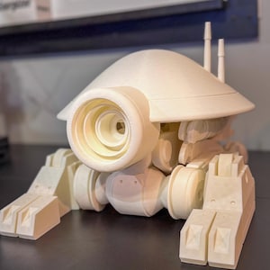 Op de afbeelding: Een crèmekleurig, 3D-geprint model van een futuristisch voertuig. Het model heeft een grote, cirkelvormige oogachtige structuur aan de voorkant, een koepelvormige bovenkant en rechthoekige voeten. Twee kleine, cilindrische antennes steken uit de bovenkant.