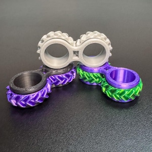 May include: A collection of 3D-printed finger rings in various colors. One ring is gray, two are purple with green accents, and one is black. The rings have a unique design, with a connecting bridge between the two ring openings.