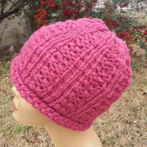 May include: A magenta-colored, hand-knitted beanie hat. The hat features a thick, textured stitch pattern and a ribbed brim. The hat is displayed on a mannequin head, showcasing its shape and design. The background includes foliage.