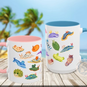 May include: Two white ceramic mugs with colorful sea slug illustrations. One mug has a pink interior and handle, the other has a blue interior and handle. The mugs are on a wooden surface with a blurred beach background.