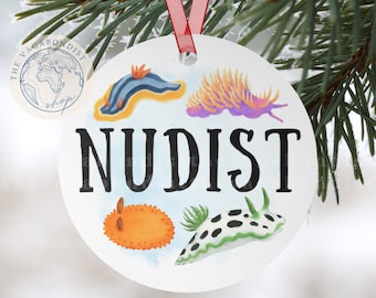 Puede incluir: Adorno navideño redondo blanco con la palabra "NUDIST" en letras negras. El adorno presenta ilustraciones coloridas de babosas marinas. Se adjunta una cinta roja para colgarlo. El adorno se muestra sobre un fondo de ramas de hoja perenne.