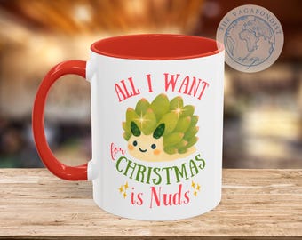 May include: White ceramic mug with a red interior and handle. The mug features a cartoon succulent with the text "ALL I WANT for CHRISTMAS is Nuds" in red and green. The mug is on a wooden surface.
