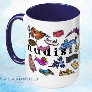 Könnte beinhalten: Weiße Keramik-Kaffeetasse mit blauem Rand und Henkel. Die Tasse zeigt eine farbenfrohe Illustration verschiedener Nacktschnecken mit dem Wort "Nudist" in schwarzen Buchstaben.