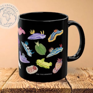 May include: Black ceramic mug featuring colorful illustrations of sea slugs. The mug has a black handle and the text "THE VAGABONDIST shop" is printed on the side. The sea slugs are in various colors and shapes.