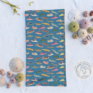May include: A rectangular fabric neck gaiter featuring a repeating pattern of colourful sea slugs on a blue background. The design includes various nudibranch species in shades of pink, yellow, orange, and brown. A circular logo reads THE VAGABONDIST shop.
