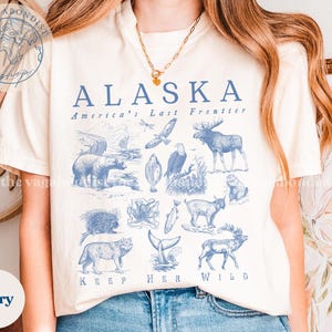 May include: Ivory-colored t-shirt with a blue graphic design featuring the word "ALASKA" and illustrations of Alaskan wildlife. The text "America's Last Frontier" and "KEEP HER WILD" are also included.