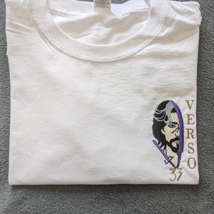 Może przedstawiać: Biała koszulka z okrągłym dekoltem i krótkimi rękawami. Koszulka posiada haftowany wzór na lewej piersi przedstawiający stylizowaną twarz z napisem "VERSO" i numerem "33" złotą nicią.
