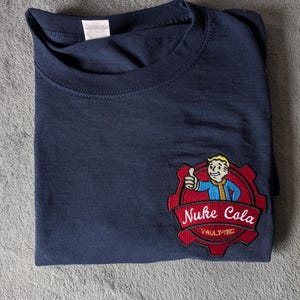 Puede incluir: Camiseta azul marino doblada con un parche bordado rojo y negro que presenta un personaje de dibujos animados y las palabras "Nuke Cola" y "Vault-Tec". La camiseta está sobre una superficie gris.