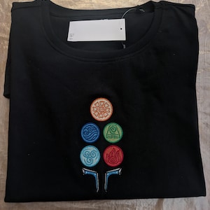 Könnte beinhalten: Ein schwarzes Kurzarm-T-Shirt mit einem gestickten Motiv auf der Brust. Das Design besteht aus fünf kreisförmigen Symbolen in Orange, Blau, Grün, Hellblau und Rot, die in einer Gruppe über zwei kleinen, symmetrischen blau-weißen Formen angeordnet sind.