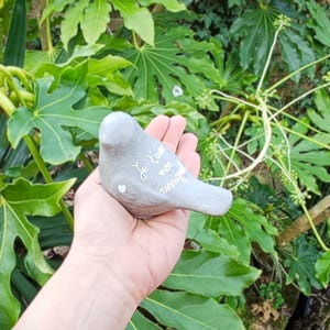 Puede incluir: Una escultura gris en forma de pájaro con el texto "Je t'aime mon rossignol" y un pequeño corazón. La escultura se sostiene en una mano sobre un fondo de follaje verde. El pájaro está hecho de un material liso y mate.
