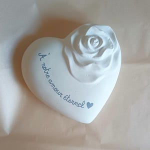 May include: A white heart-shaped ornament featuring a raised rose and the inscription "À notre amour éternel" in light blue. A small blue heart is also present. The object rests on a light brown surface.