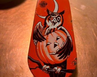 May include: Vintage Halloween toy with an owl perched on a jack-o'-lantern. The orange and white graphic is set against a red background. The toy is made of metal and has a slightly worn appearance, adding to its vintage charm.