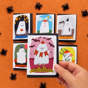 May include: Five rectangular greeting cards featuring white ghost illustrations with Halloween themes. The front card shows a ghost holding a banner that reads BOO, wearing a pumpkin hat and striped socks, with bats in a pink sky and Happy Halloween text.