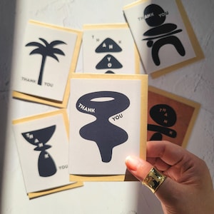 Puede incluir: Un conjunto de tarjetas de agradecimiento con diseños abstractos negros sobre fondos blancos. Cada tarjeta presenta las palabras "THANK YOU" en una fuente sencilla. Las tarjetas se muestran con sus sobres de papel kraft.