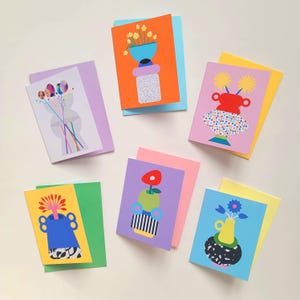 Puede incluir: Seis tarjetas de felicitación con diseños florales abstractos y coloridos. Cada tarjeta presenta una ilustración única de flores en jarrones o arreglos, sobre fondos vibrantes de color naranja, azul, amarillo y morado. Las tarjetas se combinan con sobres de colores a juego o complementarios.