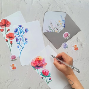 Puede incluir: Juego de papelería blanco con diseños florales. Incluye sobres, tarjetas y pegatinas. Las tarjetas presentan ilustraciones de acuarela de amapolas rojas, flores azules y peonías rosas. Una mano escribe en una tarjeta.