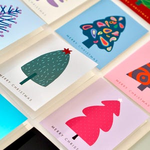 May include: A collection of rectangular greeting cards featuring various stylised Christmas tree illustrations in colours including purple, blue, green, pink, and black, each printed with the text Merry Christmas in a simple sans-serif font.