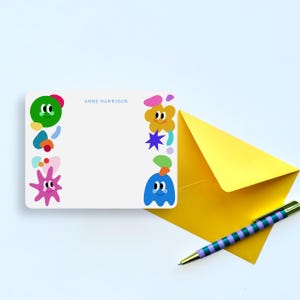 Pode incluir: Um cartão de papelaria retangular branco personalizado com o nome ANNE HARRISON em texto azul. O cartão apresenta formas coloridas estilo desenho animado com rostos nas bordas, acompanhado por um envelope amarelo brilhante e uma caneta listrada.