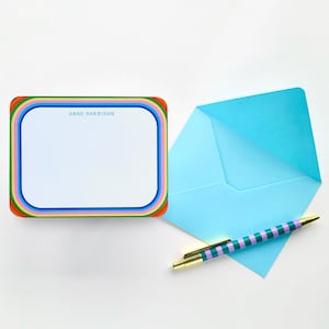 Peut inclure: Une carte de correspondance rectangulaire blanche avec une bordure arc-en-ciel multicolore et le nom ANNE HARRISON imprimé en bleu. À côté se trouvent une enveloppe bleu clair et un stylo à rayures violettes et vertes avec un clip doré.