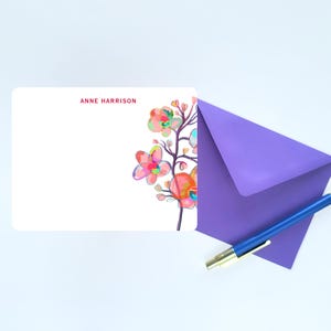 May include: A white rectangular stationery card with the name ANNE HARRISON printed in red at the top left. The right side features a colourful watercolour-style floral illustration. A purple envelope and a blue ballpoint pen are positioned nearby.