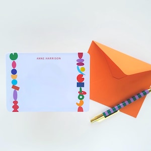May include: A white rectangular stationery card with the name ANNE HARRISON printed in red, featuring vertical borders of colourful geometric shapes. An orange envelope and a pen with green and purple stripes are positioned next to the card.